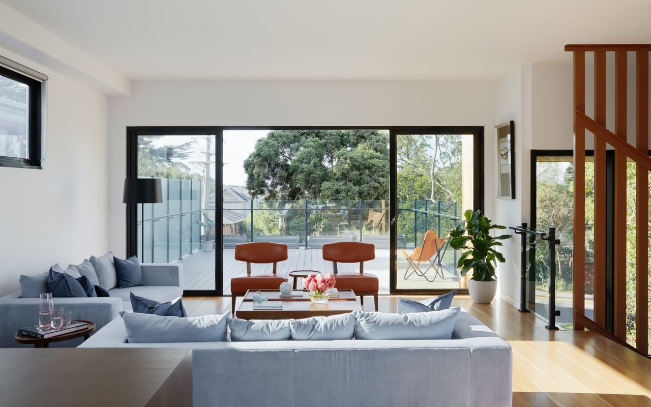 A living room with light blue sofas, brown leather chairs, and large sliding doors leading to a balcony with trees.